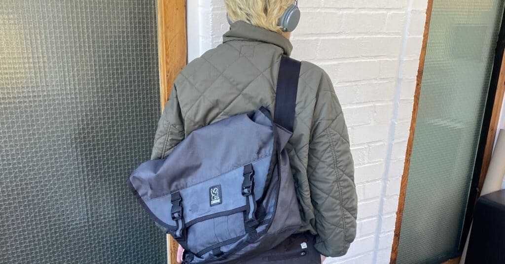 a collegiate messenger bags for school is a Chrome bag worn by a person with blond hair and an olive green bomber jacket
